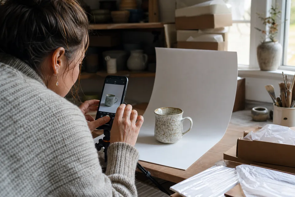 Seller photographing a ceramic mug on a white sweep near a window