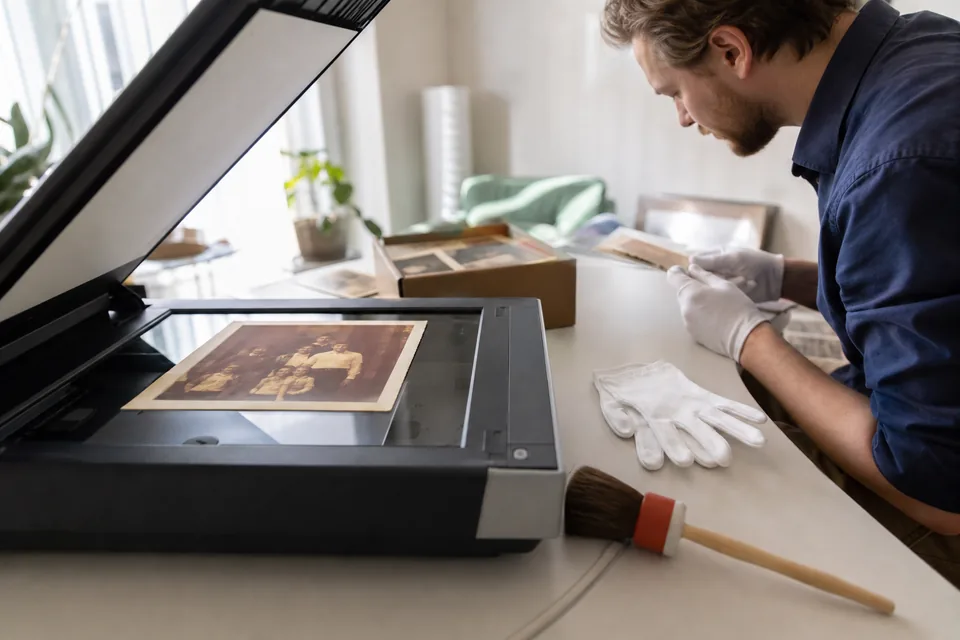 Old family prints laid on a flatbed scanner beside cotton gloves